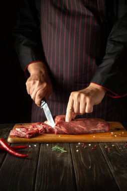Slicing raw beef meat by chef hands on kitchen cutting board. Idea for a hotel menu on a dark background.