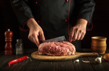 The chef with a knife in his hand in the kitchen prepares to cut raw meat before baking or barbecue. Menu for a restaurant or hotel
