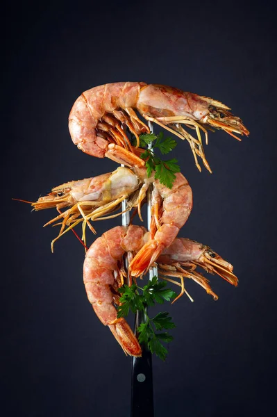 Three boiled tiger prawns or king shrimps with parsley on a fork. Delicious street food idea on black background.