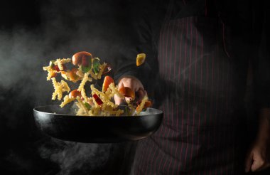 Cooking Thai food in a restaurant with fresh vegetables and pasta with spices. The chef throws food on a hot pan. Space for advertising on a black background