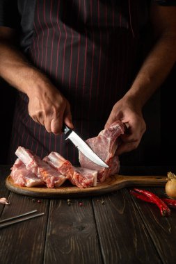 Slicing raw ribs with butcher or cook hands on kitchen cutting board. Barbecue grill. Idea for a restaurant or hotel menu on a dark background