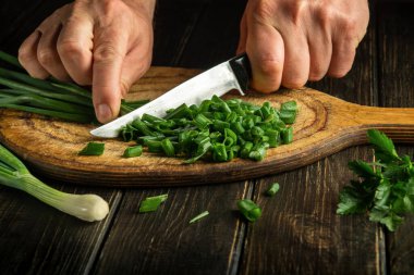 The chef cuts with a knife green onion on a cutting board for preparing a vegetarian dish.