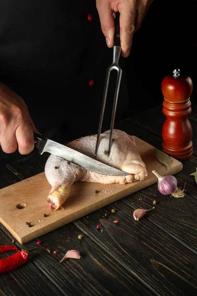 The cook or chef cuts a fresh raw chicken drumstick on a dark background. Nearby on the kitchen table are ingredients for cooking and spices.