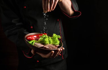 Professional chef sprinkles salt on a sliced steak with beef and vegetables in a plate. The concept of serving dishes to order