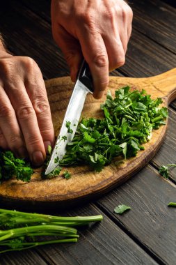 Cook hands cutting green parsley leaves on a cutting board with a knife for preparing a vegetarian dish.