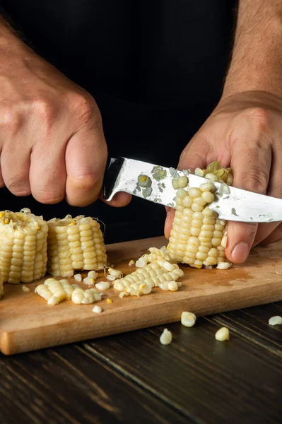 Cooking corn or maize grain by the hands of a chef with a knife on a wooden cutting board. Vegetarian food concept with advertising space
