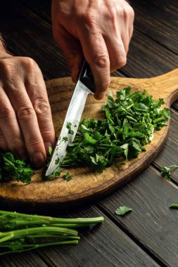 The cook hands cutting green parsley leaves on a cutting board with a knife for preparing a vegetarian dish. 