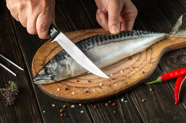 Fish chef cuts mackerel or scomber with knife on kitchen cutting board before cooking with spices and pepper.