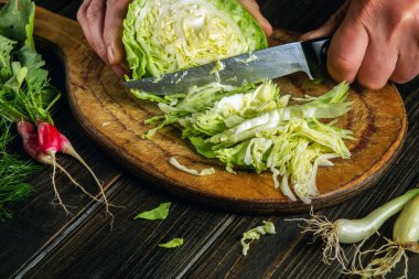 The cook cuts fresh cabbage with knife for salad on a kitchen table with fresh vegetables. Cooking and restaurant or hotel concept