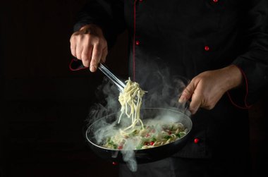 Aşçı sıcak tavada baharatlı ve baharatlı spagetti kızartıyor. Siyah arka planda reklam için yer var. İtalyan ulusal yemeği.