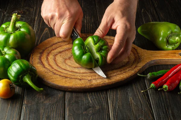 The chef cuts fresh green peppers on a wooden cutting board. Close-up of cook hands while preparing vegetarian food