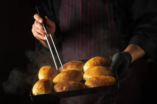 The cook bakes pies or buns on a hot baking sheet taken out of the oven. Checking readiness with a fork in the chef hand. Black space for menu or recipe