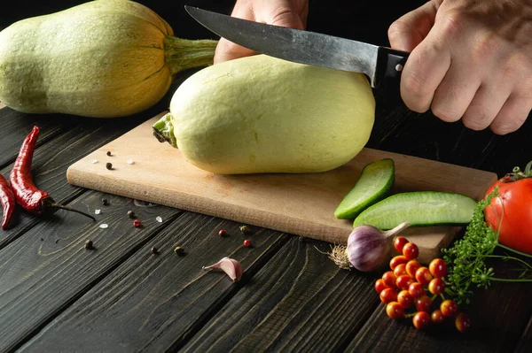 Slicing vegetable marrow with a knife before cooking by the hands of the cook on a wooden cutting board. In cooking, the thick flesh of zucchini is used