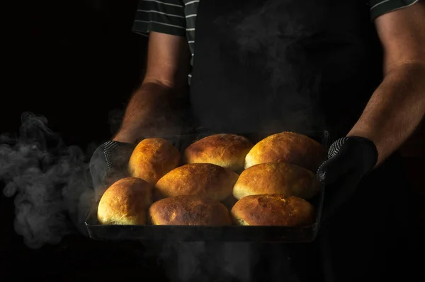 The cook holds a baking sheet with freshly baked buns or pies. Black space for menu or recipe