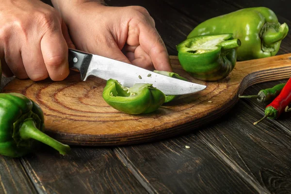 The cook cuts fresh green peppers on a wooden cutting board. Close-up of chef hands while preparing vegetarian food.