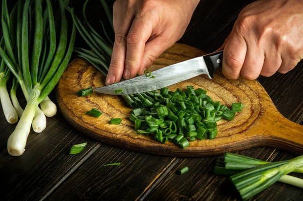 The hands of the cook with a knife cut young green onions on a cutting board. Peasant dish.