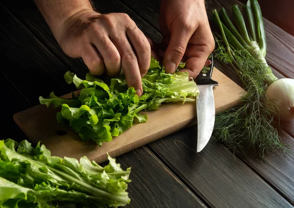 Professional cook prepares a salad on a kitchen cutting board. Close-up of chef hands on the table while preparing food.