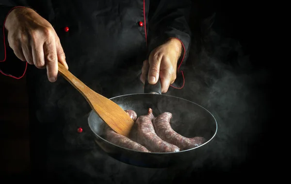 The cook frying sausages for hot dogs in a frying pan. Close-up of a chef hands with a hot pan in the kitchen. Free space for advertising on black background