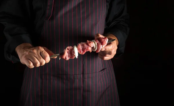 Professional chef is holding a shish kebab with raw lamb meat and onions on a skewer