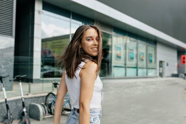 Siyah saçlı, parlak makyajlı, şık bir kadının arka portresi. Kameraya bakıyor ve şehirde yürüyor.