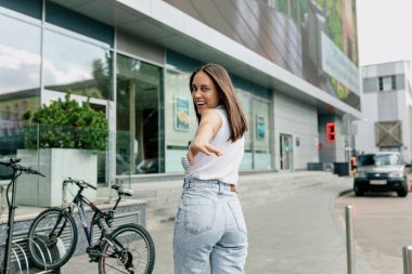 Siyah saçlı, beyaz tişörtlü, kot pantolonlu, gülümseyen ve kameraya el sallayan sevimli bir kız. 