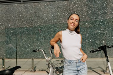 Parlak makyajlı, rahat beyaz tişört ve kot pantolon giyen, gülümseyen ve şehirde motosikletlerin yanında poz veren kendine güvenen güzel bir kadın.