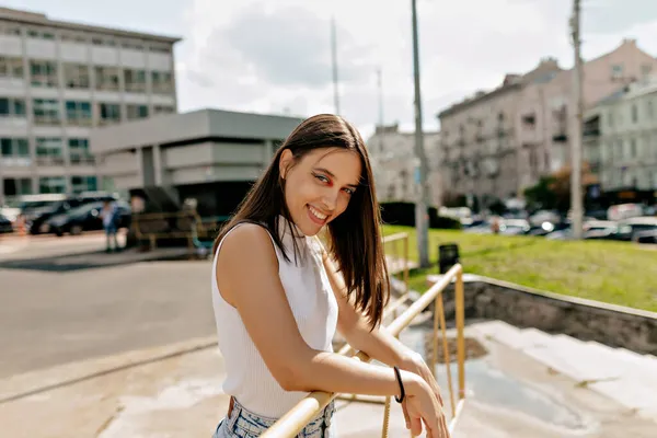 Wanita yang gembira dengan liburan yang cerah berpose dan tersenyum di depan kamera di bawah sinar matahari di latar belakang kota besar — Stok Foto