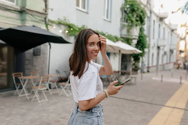 Pretty adorable lady with brown hair turn around at camera and posing at camera while walking on the street and using smartphone — Stok Foto