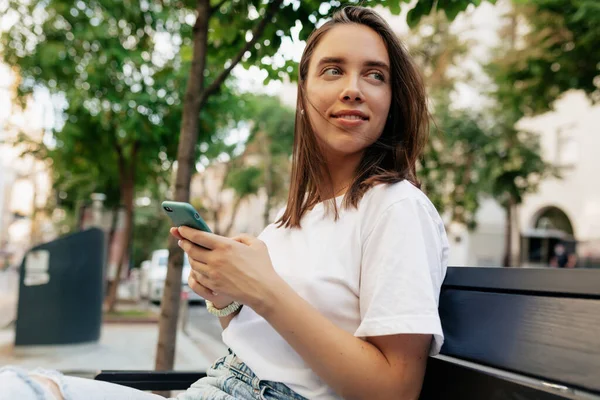 Portrait of lovable brunette young woman sitting on bench in park and using smartphone. Smiles, looks happy and enjoy life. — Stok Foto