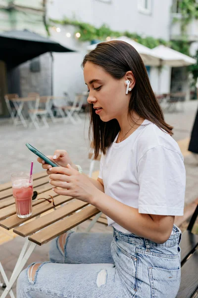 Close up outdoor portrait of stylish woman in white t-shirt using smartphone and listening music while resting in open air terrace — Stok Foto
