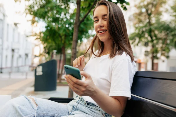 หญิงสาวชาวยุโรปที่มีความสุขนั่งอยู่บนถนนยิ้มกว้างถือสมาร์ทโฟนและมองกล้อง นางแบบวัยรุ่นพักผ่อนกลางแจ้งในเมือง . — ภาพถ่ายสต็อก