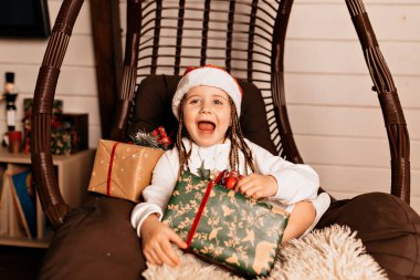 Excited little charming girl in white dress and Santa cap laughing and opening present in big wood chair. Holiday portrait of adorable lovely kid