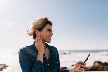 Saçlarını dalgalandıran sevimli sarışın kadın kulaklığına dokunuyor ve okyanusun arka planında akıllı telefon tutuyor ve kumsalda kayalar var. Yüksek kalite fotoğraf