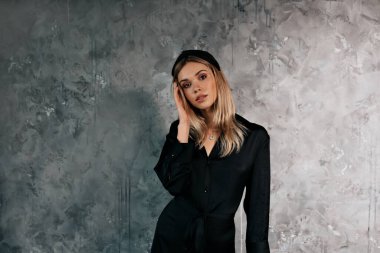 Moda kıyafetleri içinde şık bir kadın gri duvarın üzerinde kameraya ışıkla poz veriyor. Çekici sarışın kız yanağına dokunuyor ve nazikçe kameraya poz veriyor. Yüksek kalite fotoğraf