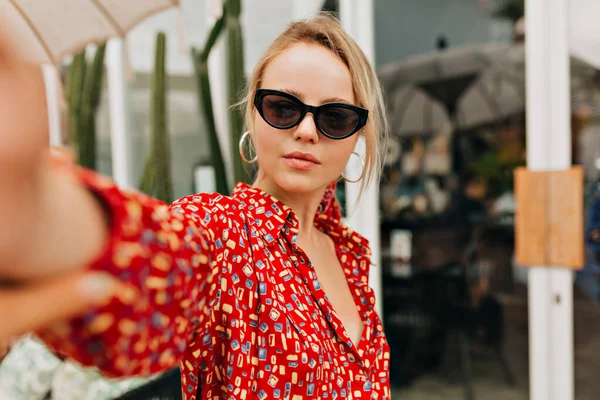 Sarışın, güneş gözlüğü takan ve parlak yaz elbisesi giyen güzel genç bir kadın şık bir kafeteryanın arka planında selfie çekiyor. — Stok fotoğraf