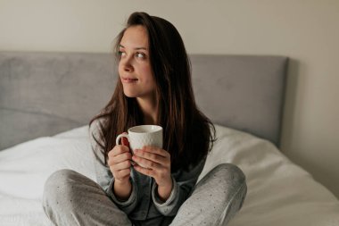 Bej hırkalı ve pantolonlu neşeli bayan yatağa oturup sabah kahvesini tutuyor. Esmer kadın yatak odasında rahatlar ve gülümser. Yüksek kalite fotoğraf