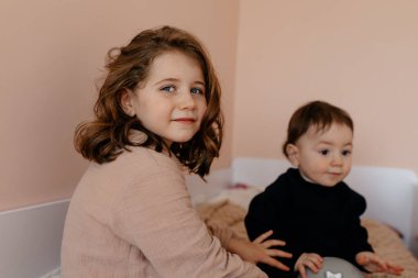 Ön planda dalgalı saç stili olan küçük sevimli kız kameraya bakıyor ve arka planda kardeşiyle gülümsüyor. Onlar sabah çocuk odasında oynuyorlar. Yüksek kalite fotoğraf