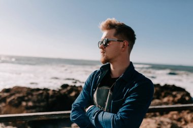 Sarı saçlı, kot gömlek giyen inanılmaz mutlu stil sahibi adam okyanusun bulanık arka planında güneş ışığı dalgalarıyla poz veriyor. Adam okyanusa ve kayalıklara karşı duruyor. Yüksek kalite fotoğraf