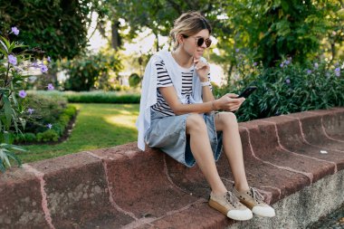 Şort giyen ve güneş gözlüklü çizgili tişörtlü genç bir kadının güneş ışığında parkta dinlenirken çekilen akıllı telefonunun tam boy portresi. Yüksek kalite fotoğraf