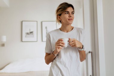 Pijamalı genç bir kadının kapalı bir portresi elinde kahve bardağıyla modern aydınlık dairenin camına bakıyor. Yüksek kalite fotoğraf