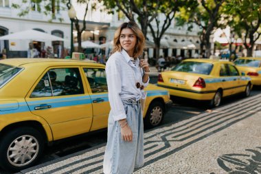 Hafif dalgalı saç stili olan şık Avrupalı kadın şehir sokaklarında egzotik ağaçlarla ve taksilerle yürüyor. Şehirdeki gezgin bir kadının açık hava portresi. Yüksek kalite fotoğraf