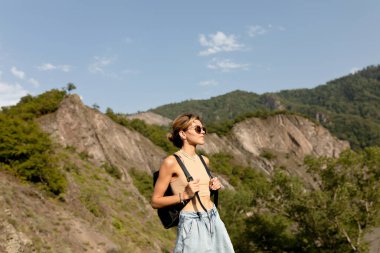 Bej tişört giyen şık hippi bayan ve sırt çantalı şort güneşli yaz gününde Avrupa dağlarında seyahat ediyor. Uzun yoldan sonra güzel bayan tepenin üstünde duruyor. Yüksek kalite fotoğraf