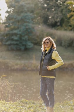 Tişört açık renk saçlı, sarı kazak ve siyah yelek giyen çekici Avrupalı kadın, ılık bahar gününde göl ve yeşil ormana karşı poz veriyor. Yüksek kalite fotoğraf