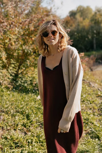 秋の森を背景に 波状の薄毛につる性のドレスとサングラスのベージュのセーターを着た素敵な魅力的な女性がカメラにポーズをとっています 高品質の写真 — ストック写真