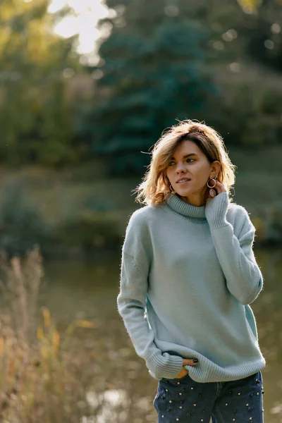 스타일의 여자가 파란색 스웨터를 호수가 보이는 공원을 걷다가 카메라 포즈를 — 스톡 사진
