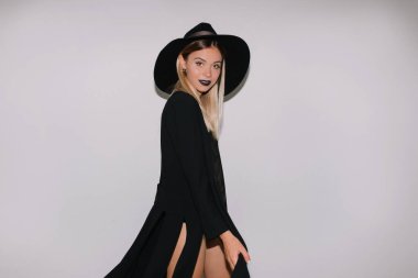 Tatil makyajlı genç bayan portresi izole edilmiş arka planda duruyor. Yakışıklı sarışın siyah elbise giyiyor ve beyaz duvara karşı siyah şapka takıyor.