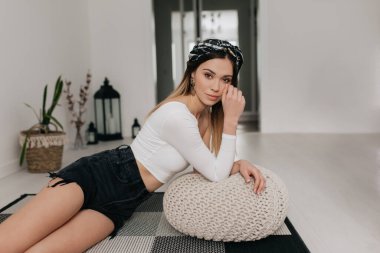 Güzel bir kadın evdeki oturma odasında yerde oturuyor, kameraya güzel bir gülümsemeyle bakıyor ve evde dinleniyor. Sarışın üstte ve şortlu.. 