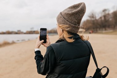 Kapüşonlu hoş bayanın arka tarafındaki portre, güneşli bir günde akıllı telefondan fotoğraf çekiyor. Kadın sahilde yürüyor ve akıllı telefon kullanıyor. Yüksek kalite fotoğraf