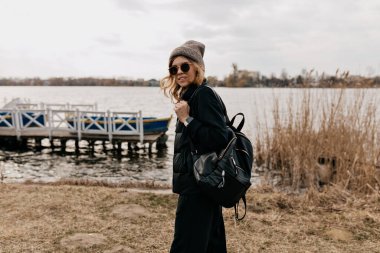 Güneş gözlüklü sırt çantalı şık bir kadın göl ve teknelere karşı kameraya poz veriyor. Kadın sırt çantasıyla seyahat ediyor. Baharda sahilde yürüyen mutlu kız. Yüksek kalite fotoğraf