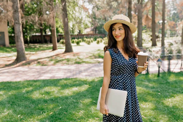 Spektakulär charmig dam med mörkt hår bär klänning och hatt håller laptop och kaffe i parken — Stockfoto